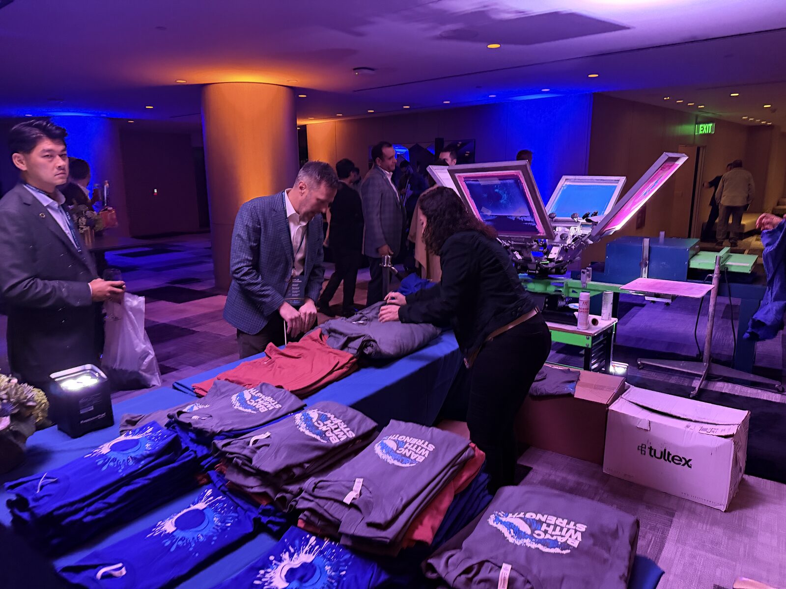 Stacks of freshly printed sponsor shirts on a table beside a live press at an evening corporate event