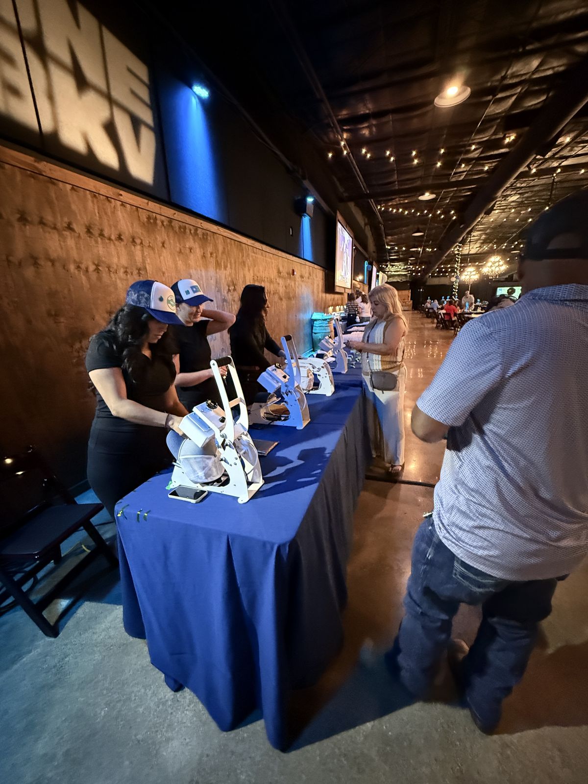 Row of hat presses staffed by crew members finishing trucker caps at an evening conference reception